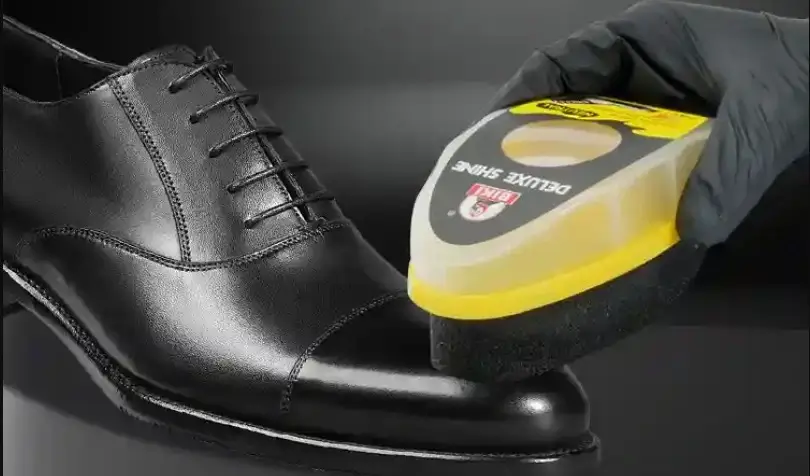 A hand wearing a black glove uses Biki Deluxe Shine sponge to polish a black leather dress shoe.